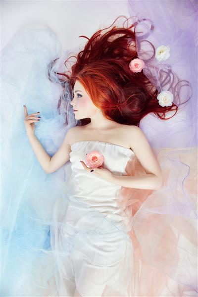 زن رمانتیک با موهای بلند و لباس ابری دختری که رویای آرایش روشن و بدن عالی دارد دختر مو قرمز با لباس رنگی روشن روی زمین پس زمینه سفید دراز کشیده است گل های زیبا در موهای دخترانه