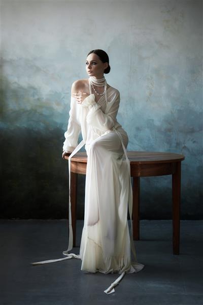 دختر سبزه باریک و زیبا که با لباس سفید بلند روی مبل نشسته است پرتره زنی با جواهرات روی گردن مدل مو و لوازم آرایشی زنانه مجموعه جدید لباس های سبک