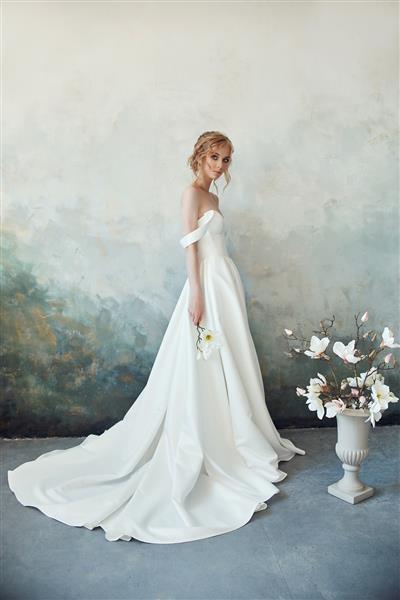 بلوند باریک و زیبا در زیر آفتاب عصر با یک لباس بلند سفید پرتره زنی با گلی در دست مدل مو و لوازم آرایشی عالی عروس مجموعه جدید لباس عروس