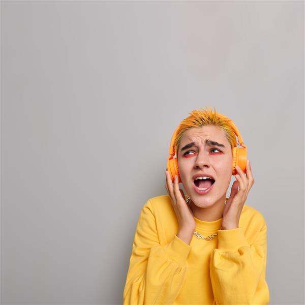 عکس عمودی از دختر شاداب با موهای زرد با ظاهری غیرعادی با آرایش روشن و با هدفون بی‌سیم به موسیقی گوش می‌دهد و در امتداد جدا از دیوار خاکستری آواز می‌خواند