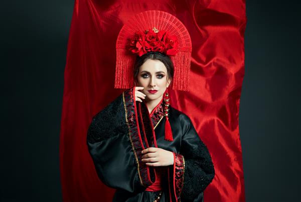 زن در لباس محلی ژاپنی قرمز چینی پوشیده است پارچه پرنده چتر و پنکه زیبا به سبک ژاپنی چینی گوشواره بلند در گوش دختری که روی پس زمینه تیره ژست گرفته است