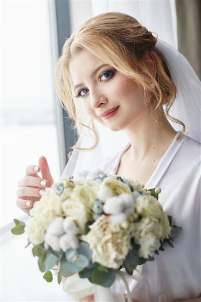 پرتره صبح عروس که برای مراسم عروسی آماده می شود پرتره زنی بلوند در نزدیکی پنجره با لباس سفید و چادری بر سر