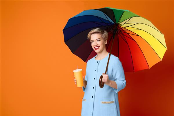 زن بلوند روشن با فنجان کاغذی قهوه زیر چتر رنگین کمانی در پس زمینه نارنجی فضای کپی