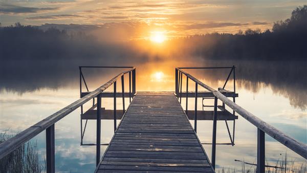 خورشید بر فراز یک دریاچه مه آلود با یک اسکله چوبی در جنگل