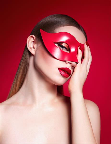 نمای نزدیک دختر جوان زیبا در استودیو در پس زمینه قرمز با ماسک قرمز روی صورت رژ لب قرمز ناخن قرمز
