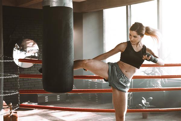زن جوان در رینگ بوکس با کیسه بوکس سنگین تمرین می کند پرتره تمرین مدل مد زیبا در سالن ورزشی لفت ضربه پا یا ضربه زانو