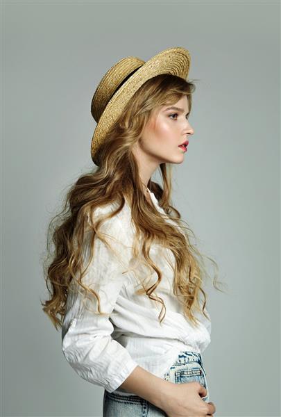 زن جوان و زیبای مو بلند با لباس‌های غیررسمی و کلاه حصیری که در استودیو روی پس‌زمینه خاکستری ژست گرفته است