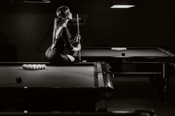 دختری با چشم بند و نشانه ای در دستانش روی میز یک باشگاه بیلیارد نشسته استبیلیارد روسی عکس سیاه و سفید