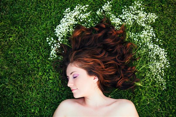 پرتره نزدیک از زن دختر جوان زیبا با موهای قهوه ای قرمز که روی چمن دراز کشیده و گل های کوچک سفید دور سرش نمای از بالای بالای سر مفهوم شادی جوانان بهار تابستان