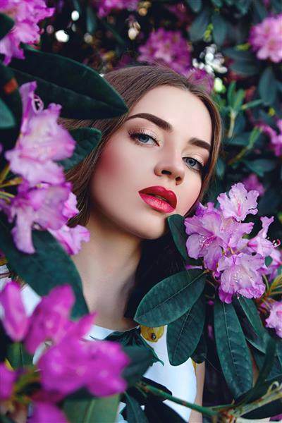 دختر زیبا در لباسی روشن با موهای روان در پس‌زمینه درختچه‌هایی با گل‌های صورتی بزرگ و برگ‌های بزرگ مد زیبایی لوازم آرایشی لوازم آرایشی طبیعی سلامت مراقبت از خود طبیعت