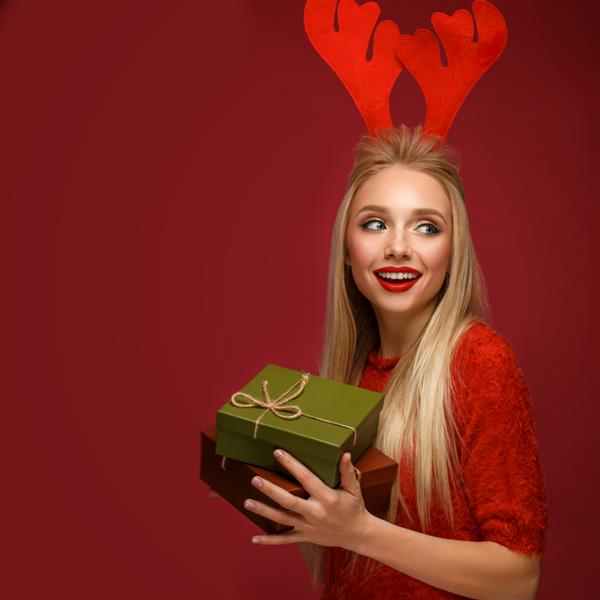دختر بلوند زیبا در تصویر سال جدید با جعبه های هدیه در دست و شاخ گوزن روی سر صورت زیبایی با آرایش جشن عکس گرفته شده در استودیو