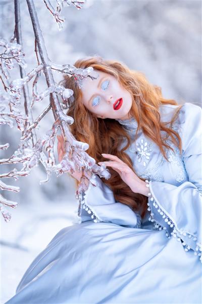 زن جوان مو قرمز یک شاهزاده خانم در یک جنگل زمستانی با لباس آبی راه می رود یخ زدگی و برف روی درختان