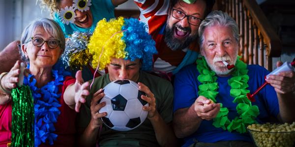 گروهی از هواداران فوتبال در خانه از موفقیت تیم لذت می برند و شادی می کنند - مفهوم تلویزیونی قهرمانی جهان 2022 - خانواده در حال تماشای فوتبال در تلویزیون در خانه و با هم در دوستی سرگرم می شوند