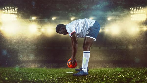 صحنه فوتبال در مسابقه شبانه با بازیکنی با لباس سفید و لباس که توپ را برای ضربه زدن قرار می دهد