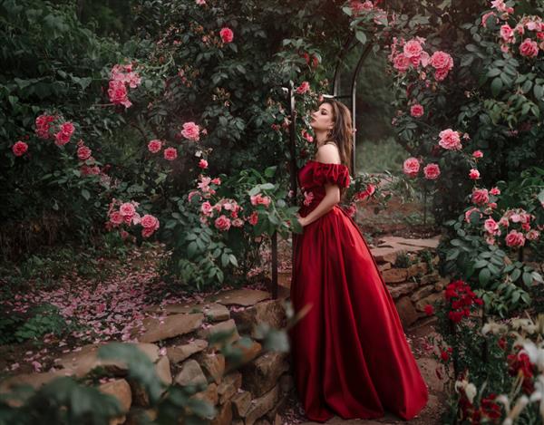 زن زیبا با لباس قرمز در حال قدم زدن در باغ پر از گل رز