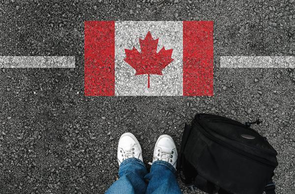 مردی با کفش و کوله پشتی روی آسفالت کنار پرچم کانادا و مرز ایستاده است