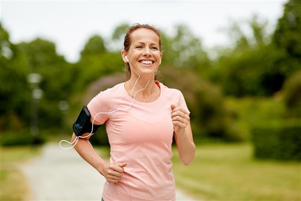 مفهوم تناسب اندام ورزش و سبک زندگی سالم - زن خندان با گوشی با بازوبند برای گوشی هوشمند دویدن در پارک تابستانی و گوش دادن به موسیقی
