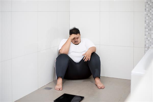 تصویر مرد چاق ناامیدی که در حمام با ترازو نشسته است که چشمانش را با دست پوشانده است