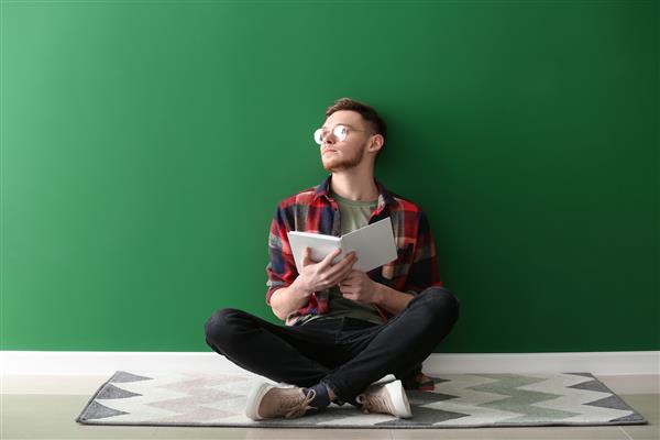 مرد جوان خوش تیپ با کتابی که نزدیک دیوار رنگی نشسته است