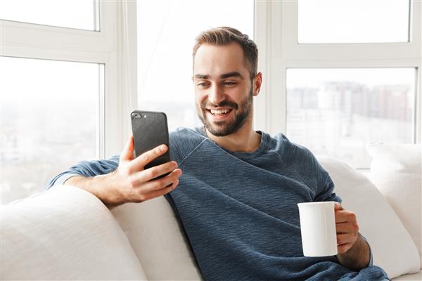 مرد جوان جذاب در حال استراحت روی مبل در خانه با استفاده از تلفن همراه نوشیدن قهوه