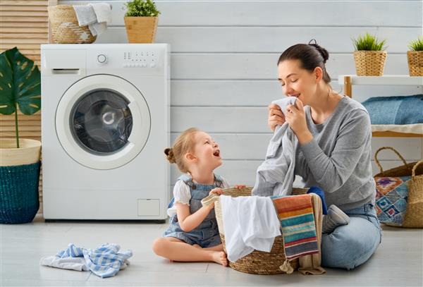 زن جوان زیبا و دختر بچه کمکی در حال سرگرمی و لبخند زدن در حال شستن لباس در خانه هستند