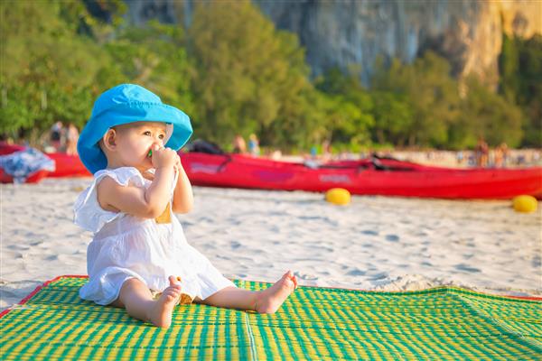 رویش دندان در 8 ماه دختر بچه ای که در ساحل نشسته و دندان گیر می جود با کلاه آبی و لباس سفید روی حصیر بامبو