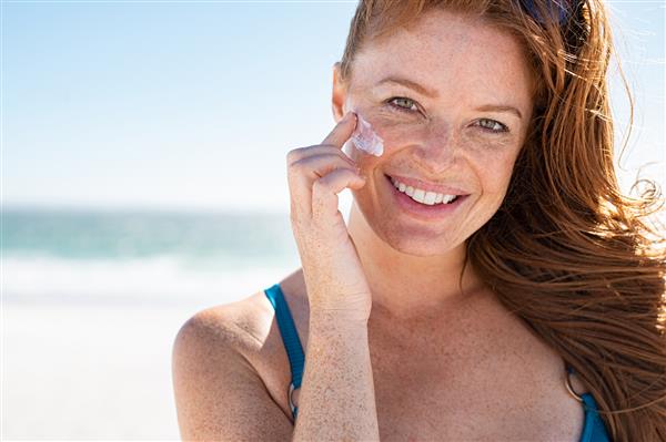 زن جوان خندان در حال استفاده از لوسیون ضد آفتاب روی صورت در ساحل با فضای کپی پرتره دختر زیبای شاد با استفاده از ضد آفتاب روی پوست ظریف خود با کک و مک و نگاه کردن به دوربین