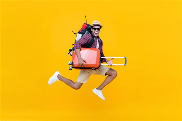 گردشگر جوان شاد آسیایی هیجان زده با چمدان پرش جدا شده روی پس زمینه زرد استودیو