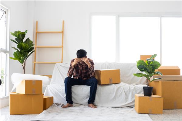 مرد مجرد آسیایی نشسته روی مبل و جعبه در آپارتمان اتاق جدید خانه و به فکر ایده خوب برای خانواده است