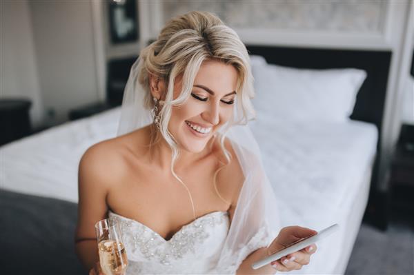یک عروس بلوند زیبا و خندان با لباس سفید روی تخت نشسته و به تلفن نگاه می کند پیام می خواند و شامپاین می نوشد پرتره عروسی یک دختر شاد