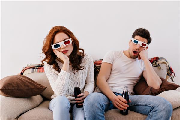 زوج بی حوصله در حال نوشیدن نوشابه روی مبل عکس داخل خانه از زن و مرد در حال تماشای فیلم سه بعدی