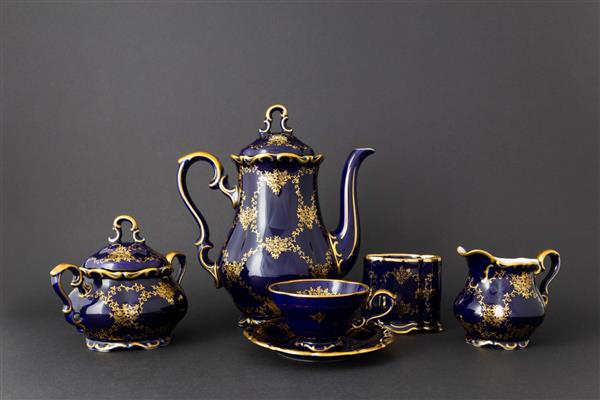 نمای نزدیک از مجموعه چای چینی وینتیج رنگ آبی کبالتی با طرح گل طلایی در زمینه خاکستری تیره این ست شامل یک قوری چای یک کاسه قند یک لیوان شیر و یک فنجان چای است