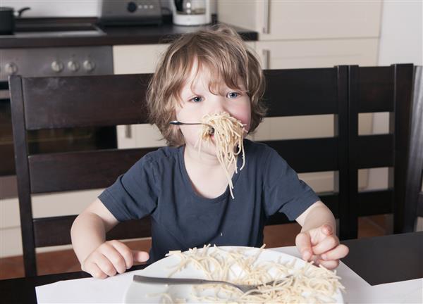 پسر جوان ناز در آشپزخانه روی میز نشسته اسپاگتی می خورد