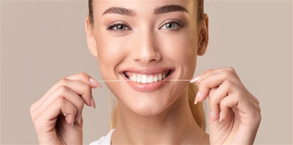 نخ دندان کشیدن زن خندان با استفاده از نخ دندان برای تمیز کردن و مراقبت از دندان‌های سفید عالی که روی پس‌زمینه بژ ظاهر می‌شود پانوراما