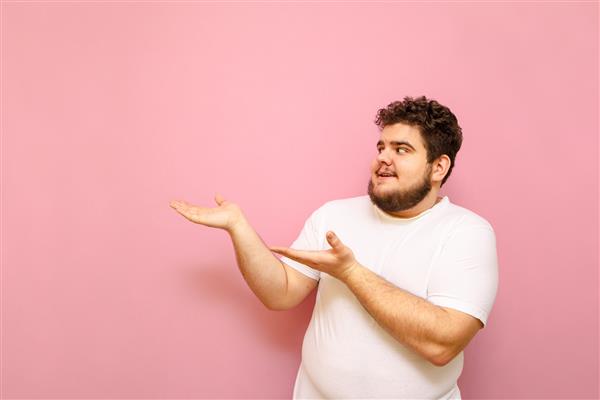 مرد جوان مثبتی که دارای اضافه وزن است روی پس‌زمینه صورتی ایستاده نگاه می‌کند و با لبخندی روی صورت دست‌هایش را به سمت فضای خالی نشان می‌دهد مرد چاق با یک تی شرت سفید دستانش را با فضای کپی تقدیم می کند