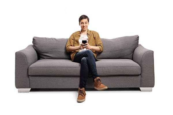 مرد جوان شادی که روی مبل خاکستری نشسته و روی یک تلفن همراه جدا شده روی پس زمینه سفید تایپ می کند