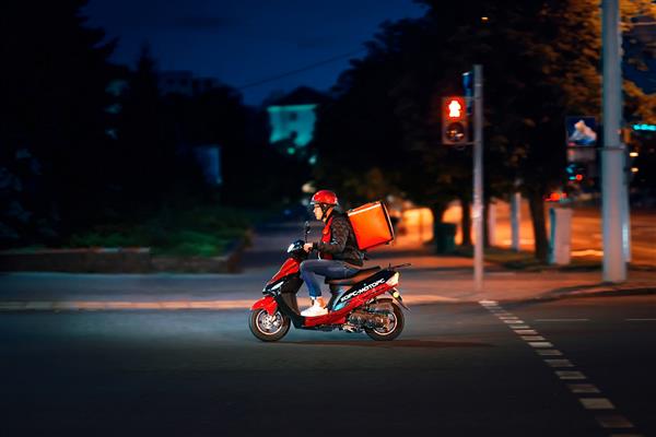 مینسک بلاروس جولای 2019 تحویل‌دهنده سفارشات آنلاین غذا را روی موتور اسکوتر در شب تحویل می‌دهد تحویل پسر جوان با کیسه حرارتی قرمز پیک رستوران غذا را حمل می کند غذا را سریع به مشتری تحویل می دهد