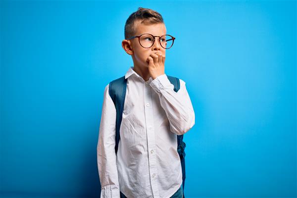 دانش‌آموز کوچک قفقازی با عینک هوشمند و کیف مدرسه روی پس‌زمینه آبی به نظر استرس‌زده و عصبی است و دست‌های روی دهانش ناخن‌هایی می‌جود مشکل اضطراب