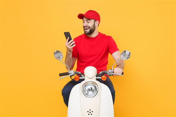 مرد تحویل‌دهنده با تی‌شرت کلاهک قرمز رانندگی با موتور سواری اسکوتر تلفن همراه جدا شده در استودیو پس‌زمینه زرد را نگه می‌دارد