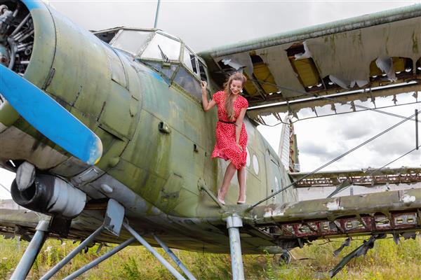 زن بالغ جوان و زیبا با لباس سنجاق قرمز روی بال هواپیمای سبز رنگ رها شده در مقابل آسمان ابری ایستاده است