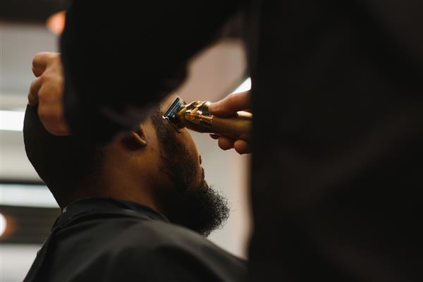 پرتره مرد جوان سیاه پوستی که با دستگاه گیره برقی حرفه ای در آرایشگاه اصلاح می شود مفهوم درمان زیبایی مردانه پسر جوان آفریقایی در حال کوتاه کردن موی جدید در آرایشگاه