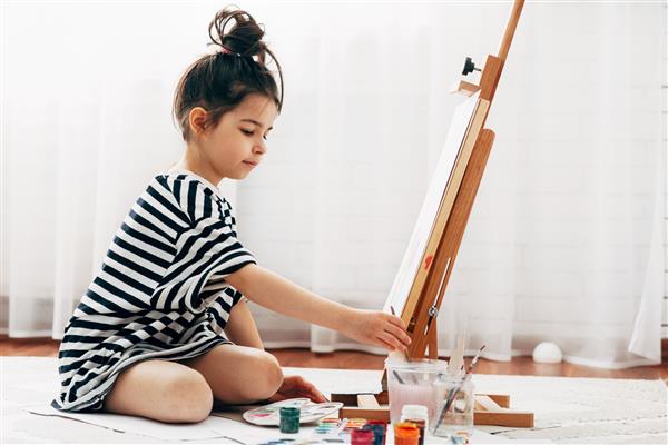 تصویر تمام قد افقی از یک بچه دختر بچه که روی کاغذ روی سه پایه در خانه نقاشی می کند یک بچه ناز روی فرش نشسته و در اتاقش نقاشی می کشد