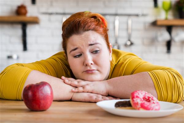 زن چاق غمگینی که بین شیرینی و میوه تغذیه سالم و غذاهای ناسالم انتخاب می کند انتخاب دو گزینه انتخاب بین خوردن دلما برای یک سبک زندگی سالم یا نه