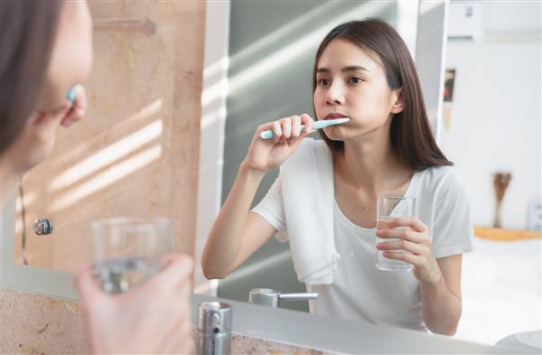 زن جوان آسیایی در حال مسواک زدن و نگاه کردن در آینه نگه داشتن لیوان آب حوله روی شانه در حمام مفهوم بهداشت دهان و مراقبت بهداشتی