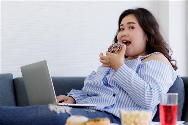 خریدار زن چاق و زیبای جوان آسیایی که در حال استراحت روی مبل پارچه ای نشسته است با استفاده از رایانه لپ تاپ به دوربین نگاه می کند و در حال جستجوی محصولات خرید آنلاین در حالی که دونات شکلاتی خوشمزه در دست می خورد