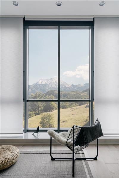 عکس عمودی از صندلی فلزی مدرن روی کف خاکستری فرش شده در مقابل پنجره بزرگ با منظره کوهستانی شگفت انگیز در اتاق نشیمن با رنگ روشن طراحی داخلی مینیمالیستی در اتاق یا خانه هتل