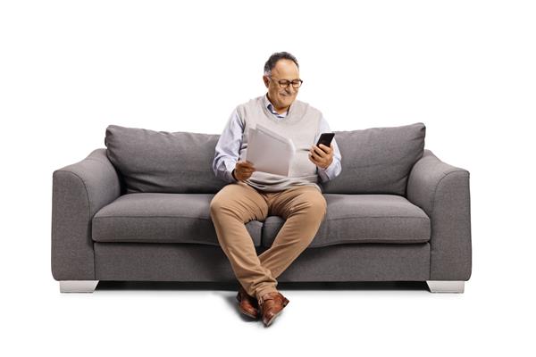 مرد بالغ با استفاده از تلفن همراه روی مبل نشسته و سند کاغذی جدا شده در پس زمینه سفید را در دست دارد