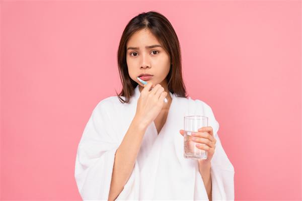 زن آسیایی با بریس هایی با مسواک زدن دندان ها و نگه داشتن لیوان آب حوله روی شانه در زمینه صورتی مفهوم بهداشت دهان و مراقبت های بهداشتی
