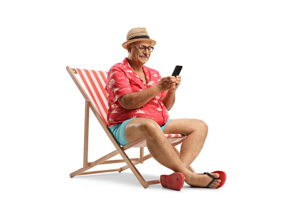 مرد سالخورده ای که روی صندلی ساحلی استراحت می کند و از تلفن هوشمند جدا شده در پس زمینه سفید استفاده می کند
