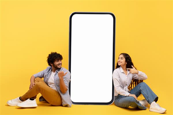 زوج هندی شادی که در نزدیکی گوشی هوشمند بزرگ با صفحه نمایش سفید خالی نشسته اند فضای کپی را برای طراحی اپلیکیشن یا تبلیغات نشان می دهند روی پس زمینه زرد ژست می گیرند بنر ماکت
